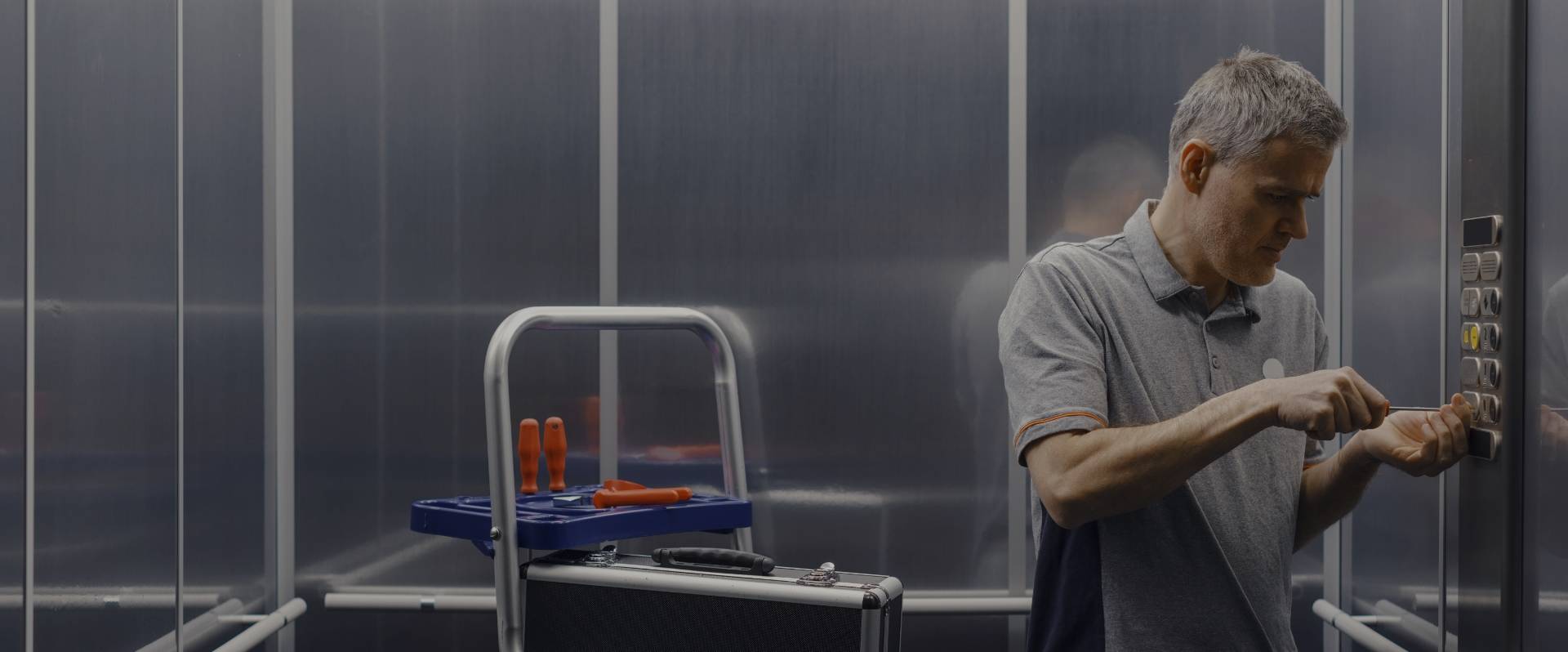 Titelbild für "Service" Handwerker mittleren Alters mit blauer Latzhose repariert einen Fahrstuhl