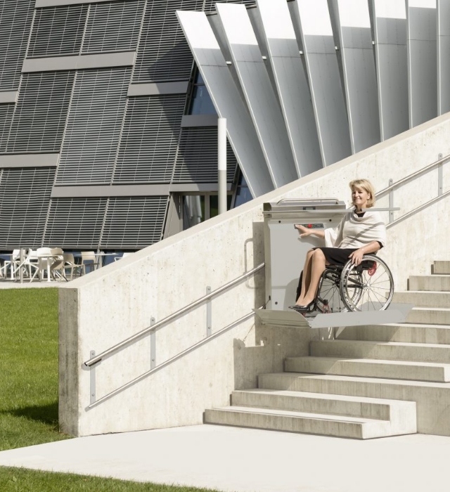 Bild zu "Plattformlift" Plattformlift im Außenbereich eines modernen Gebäudekomplexes mit einer Frau im Rollstuhl darauf