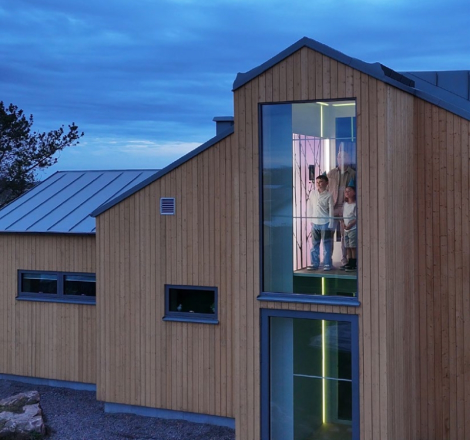 Blick von außen in ein modernes Holzhaus mit einem gläsernen Fahrstuhl, darin ein Erwachsener und zwei Kinder